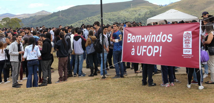Multidão de estudantes reunida ao ar livre em um evento universitário. À direita, faixa vermelha com o texto “Bem-vindos à UFOP! Universidade Federal de Ouro Preto”, com montanhas e tendas ao fundo.