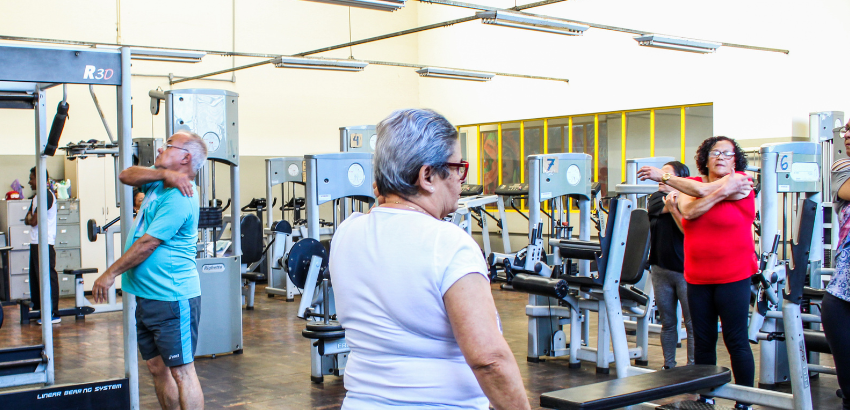 Mulher idosa de cabelos grisalhos curtos, usando camiseta branca, vista de costas em uma academia com aparelhos de musculação.