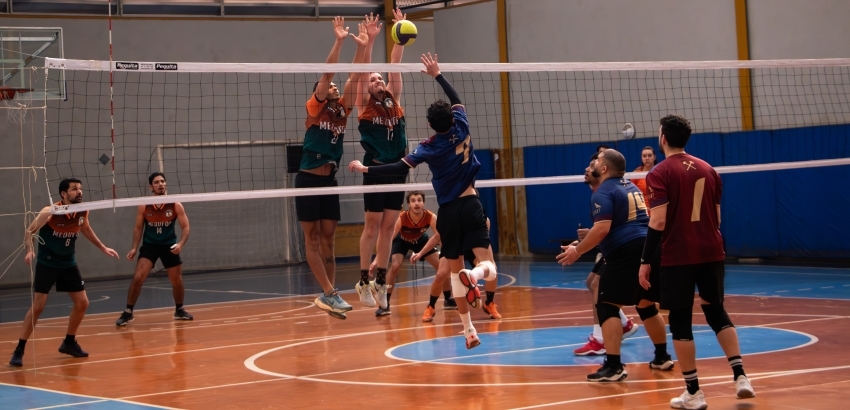 Jogo de vôlei masculino em quadra coberta. Atacante de uniforme azul salta para a cortada enquanto dois jogadores adversários, de uniforme verde e laranja, sobem para o bloqueio na rede. Outros atletas observam atentos a jogada.