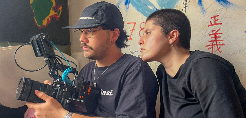 Duas pessoas observam atentamente o visor de uma câmera de cinema digital; uma segura o equipamento, enquanto a outra se inclina para ver o enquadramento. Atrás deles, parede com desenhos, rabiscos e escrita vermelha em japonês. Luz natural suave.