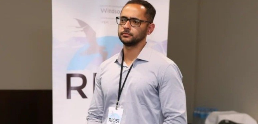 Professor Lucas Assis Attílio, no centro da imagem. Ao fundo, um banner branco.