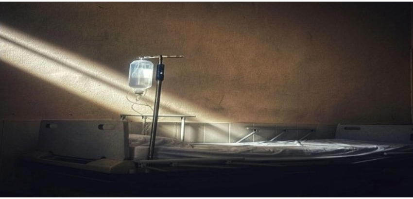 Leito hospitalar vazio com suporte de soro iluminado por um feixe de luz em ambiente escuro.