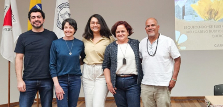 Cinco representantes da UFOP, em pé lado a lado, em ambiente interno com bandeiras ao fundo. 