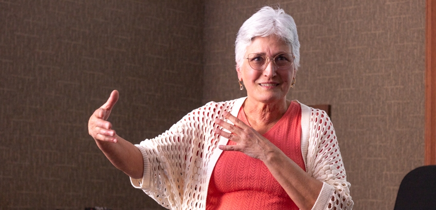 Pessoa de cabelos curtos e brancos, usando óculos, blusa coral e casaquinho branco. Em ambiente interno, gesticula com uma mão estendida e a outra sobre o peito, transmitindo expressividade e envolvimento na fala.