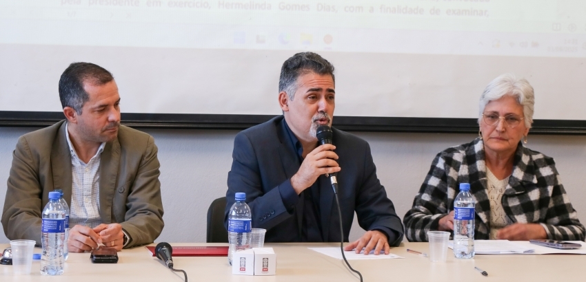 Três pessoas sentadas à mesa durante uma conferência. O homem ao centro fala ao microfone, enquanto os outros dois escutam. Há garrafas de água, copos e papéis sobre a mesa. Uma tela de projeção está ao fundo.