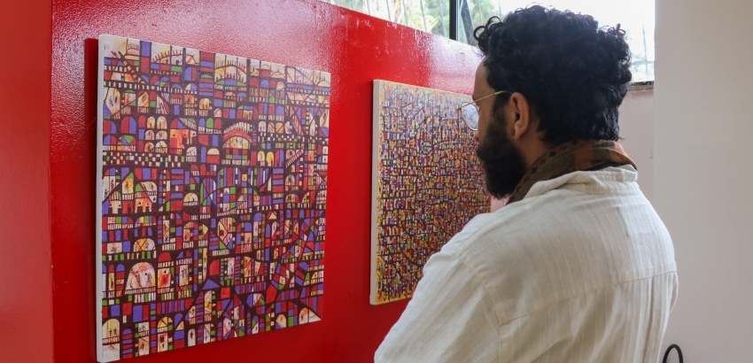 Um homem com barba e óculos, de camisa clara e cachecol, está de costas olhando atentamente para duas pinturas abstratas vibrantes, ricas em detalhes e cores, penduradas em uma parede vermelha.