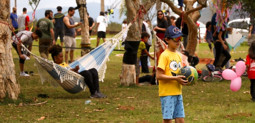 Um menino de óculos de sol, boné azul e camiseta amarela segura uma bola de futebol em um parque gramado, rodeado por árvores. Ao fundo, diversas pessoas interagem, e uma mulher relaxa em uma rede listrada.