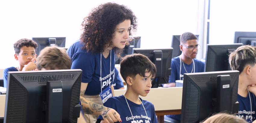 Professora orienta aluno em frente a computador em sala de informática, durante atividade do Encontro Regional PIC OBMEP. Outros estudantes, com camisetas azuis do evento, também participam concentrados ao fundo.