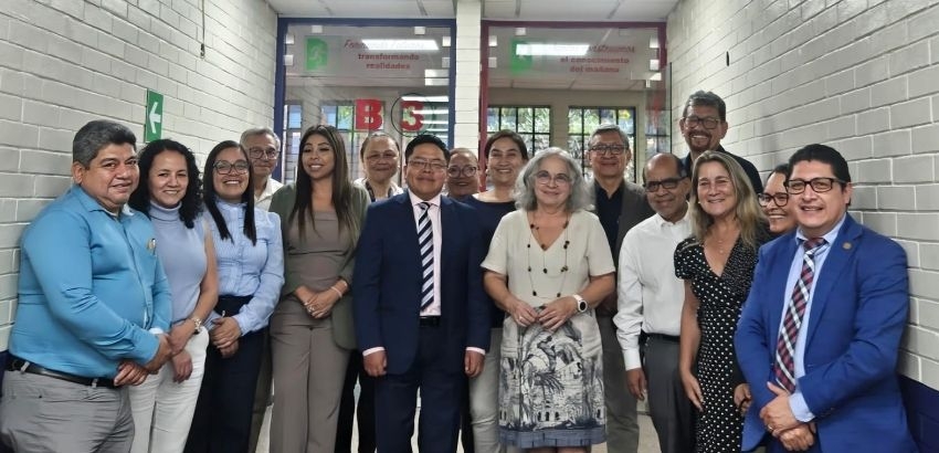Grupo de 16 pessoas posando em um corredor interno com paredes brancas. Estão alinhadas lado a lado, sorrindo para a câmera, vestindo roupas formais e casuais.
