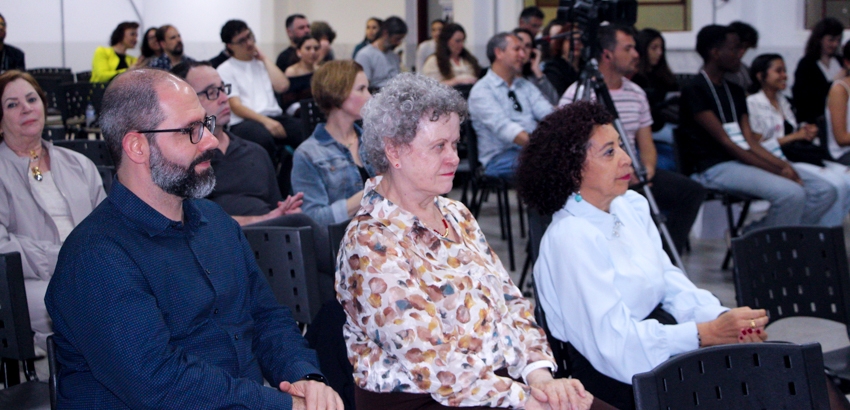 Pessoas sentadas em cadeiras assistem atentamente a um evento em ambiente fechado. Em destaque, três pessoas na frente, sendo duas mulheres e um homem, com outras pessoas ao fundo.