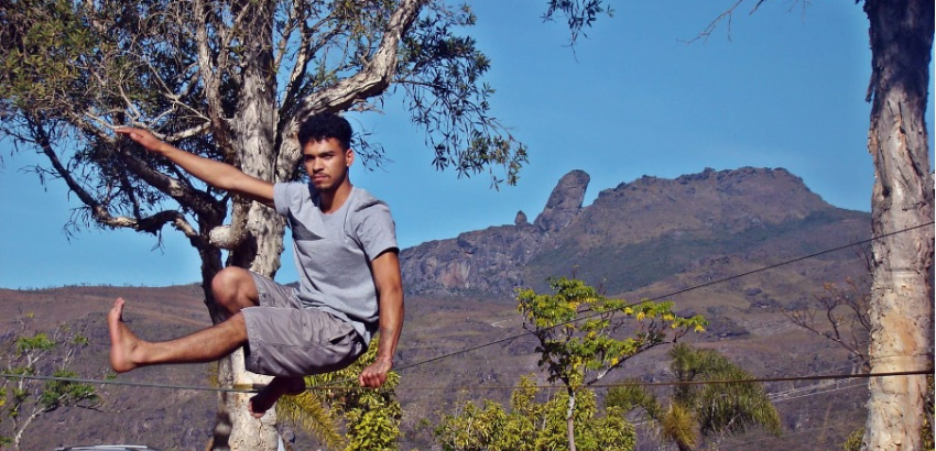Jovem pratica slackline em área externa, equilibrando-se na fita com uma perna dobrada e outra estendida. Ao fundo, montanhas e céu azul compõem a paisagem, com árvores em primeiro plano.