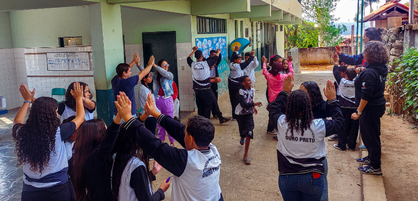 Na imagem, estudantes uniformizados participam de uma atividade em grupo ao ar livre em uma escola de Ouro Preto. Eles estão dispostos em círculo com três participantes cada, com as mãos levantadas, interagindo entre si.