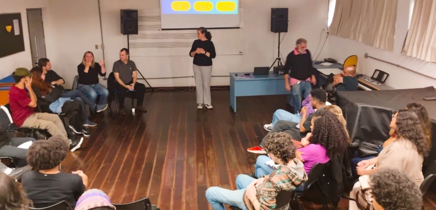 Sala de aula do DEMUS com cerca de 18 pessoas sentadas em semicírculo, duas pessoas de pé conduzindo a atividade. Piso de madeira, projetor ligado na parede, mesa azul com notebook e equipamentos, cortinas claras e piano coberto ao fundo.