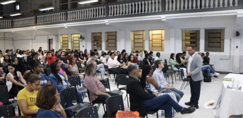 Foto mostra o deputado Leleco Pimentel segurando um microfone durante sua fala. Estantes sentados e o diretor e a vice-diretora, Harrison e Cristiane, junto com o representante da reitoria Rondon Marque na primeira fileira 