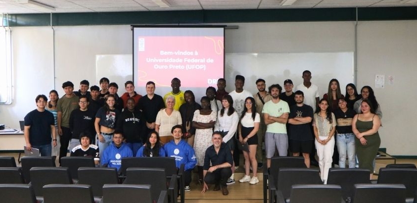 Grupo de estudantes reunidos em uma sala de aula da Universidade Federal de Ouro Preto, posando para foto em frente a um projetor que exibe uma mensagem de boas-vindas. As pessoas estão organizadas em filas, algumas em pé e outras agachadas, com cadeiras 