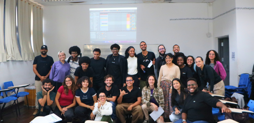 Fotografia de grupo tirada em uma sala de aula universitária. O grupo é majoritariamente composto por jovens adultos, muitos sorrindo e fazendo gestos descontraídos. Ao fundo, há uma projeção ligada em uma parede branca, exibindo a interface de um softwar