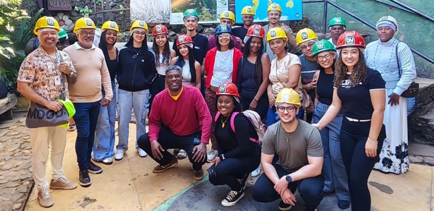 Grupo de visitantes com capacetes de segurança posa para foto durante visita à mina de ouro em Ouro Preto, em área de acesso com estruturas de pedra.