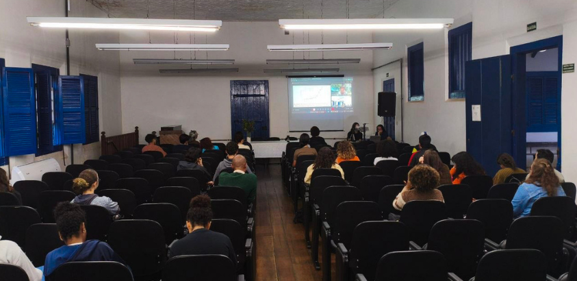 Imagem mostra um auditório com estudantes sentados e algumas cadeiras vazias. Os participantes do acompanham a apresentação via meet com professor assistente Manoel Horta Ribeiro, da Universidade de Princeton, EUA. O debate foi sobre mediação algorítimica