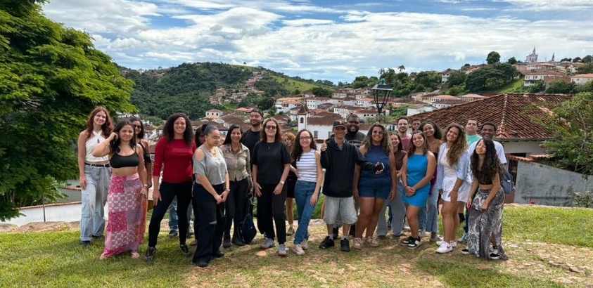 “Grupo de cerca de vinte jovens posando para uma foto em uma área gramada, com casas históricas de telhado vermelho e colinas verdes ao fundo, sob um céu parcialmente nublado.”