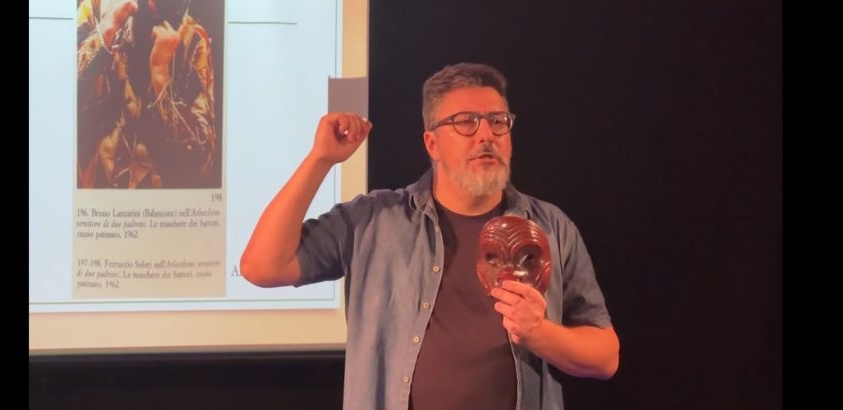 Homem de óculos, barba grisalha e camisa azul aberta sobre camiseta escura faz uma apresentação em um palco. Ele segura uma máscara marrom esculpida em uma das mãos e gesticula com a outra enquanto fala. Ao fundo, há uma tela de projeção ex