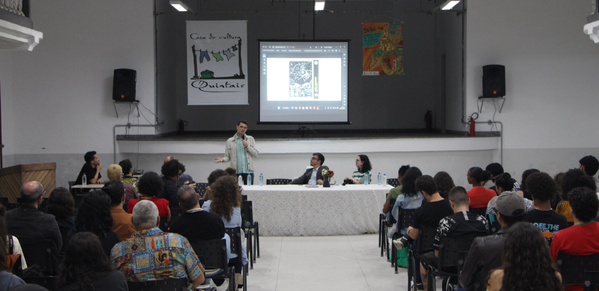 Imagem mostra o professo e jornalista, João Marcos em pé com microfone na mão, estudantes sentados e de consta e ao fundo banner do grupo quintais durante o primeiro simpósio nacional de jornalismo de quadrinhos, na UFOP, em Mariana. 