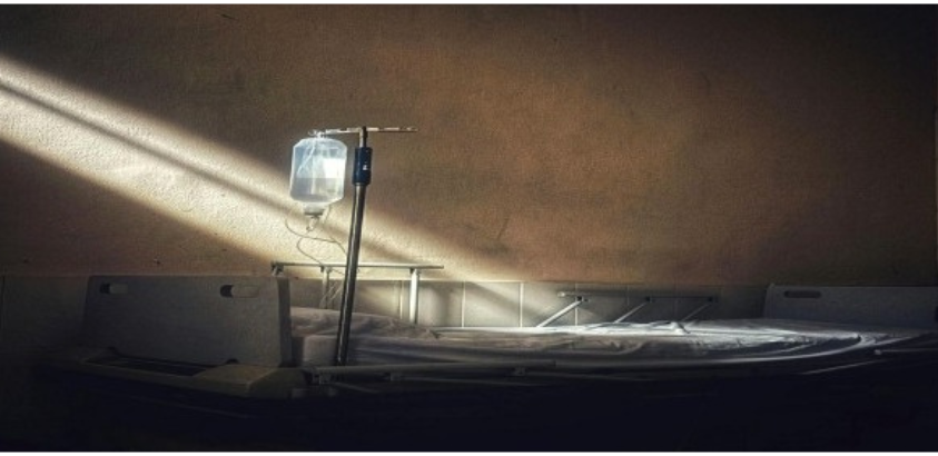 Leito hospitalar vazio com suporte de soro iluminado por um feixe de luz em ambiente escuro.