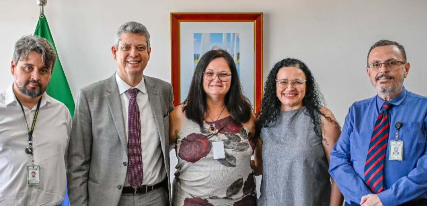 Cinco pessoas posam lado a lado em ambiente interno, com bandeira verde à esquerda e quadro central na parede. Três homens de crachá usam terno ou camisa social; duas mulheres ao centro sorriem, uma de vestido floral e outra de vestido cinza.