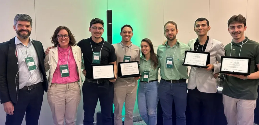 Grupo de sete pessoas posa sorridente em ambiente interno iluminado com luz verde ao fundo. Quatro seguram certificados de premiação. Todos usam crachás do evento. Vestem roupas sociais e semiformais em tons claros e escuros.