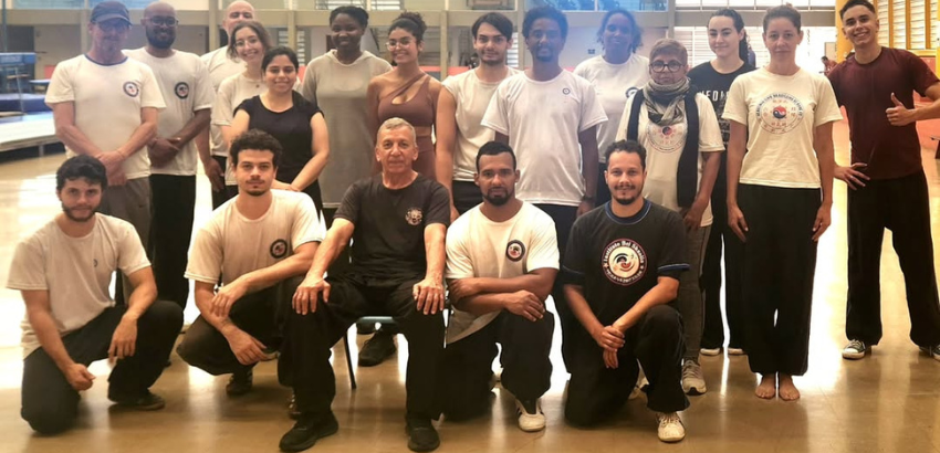 Grupo de praticantes de Tai Chi Chuan posa para foto em ambiente interno amplo e bem iluminado. As pessoas usam roupas esportivas, muitas com camisetas do grupo. Ao fundo, é possível ver tatames e equipamentos de treino.