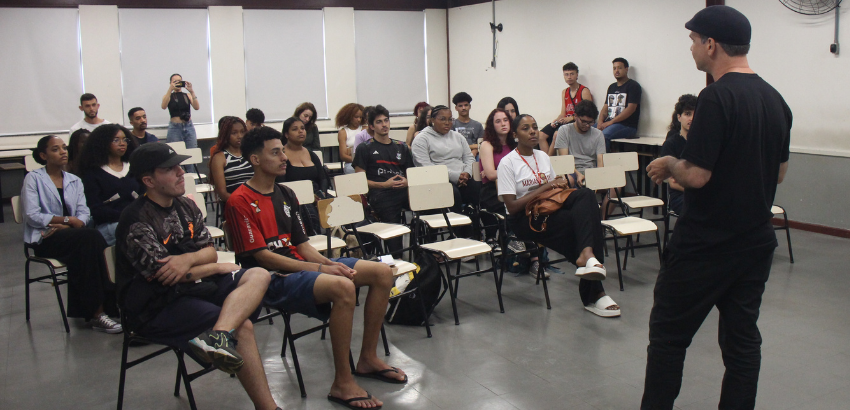 Imagem mostra estudantes em diálogo o professor Marlon Garcia da Silva. Os alunos estão sentados em cadeiras, uma aluna ao fundo está de pé e segura um celular na mão. Marlon está de costas, com roupa preta e boina. A sala é um espaço amplo e claro. 
