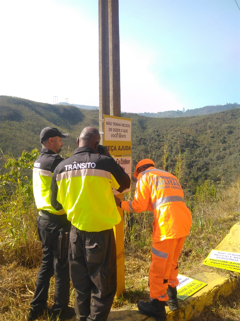 Três homens trajando uniformes da OuroTrans e Corpo de bombeiros; em cima de uma calçada, instalando uma placa em um poste; a paisagem é composta por mata e céu azul