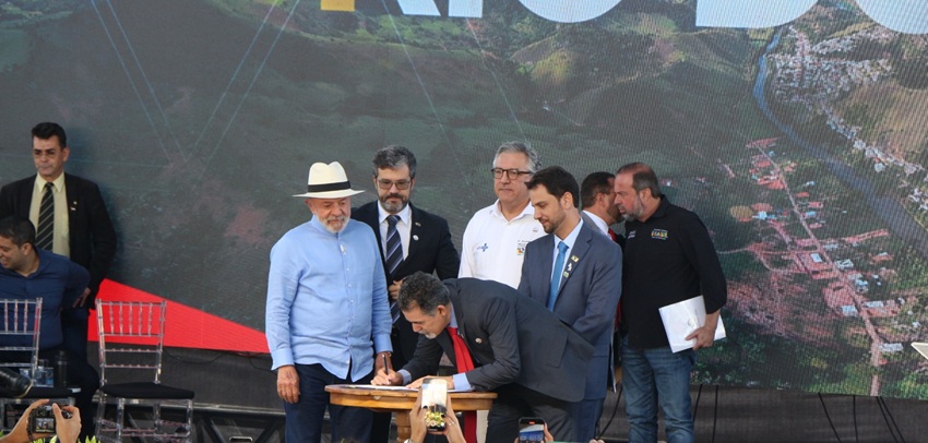 homem de terno assina documento em mesa de madeira, cercado por outros homens, incluindo um de chapéu branco. Ao fundo, imagem aérea de cidade projetada em telão e presença de seguranças e público.